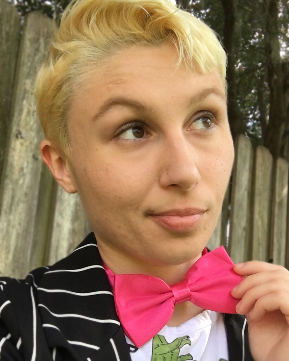 An blonde androgynous person looks off-camera wearing a pink bowtie & black & white striped blazer